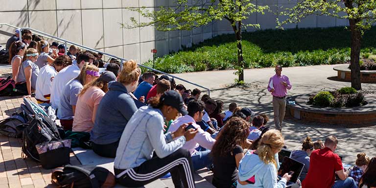 Students sitting on steps outdoors listen to a speaker who is standing under a tree.